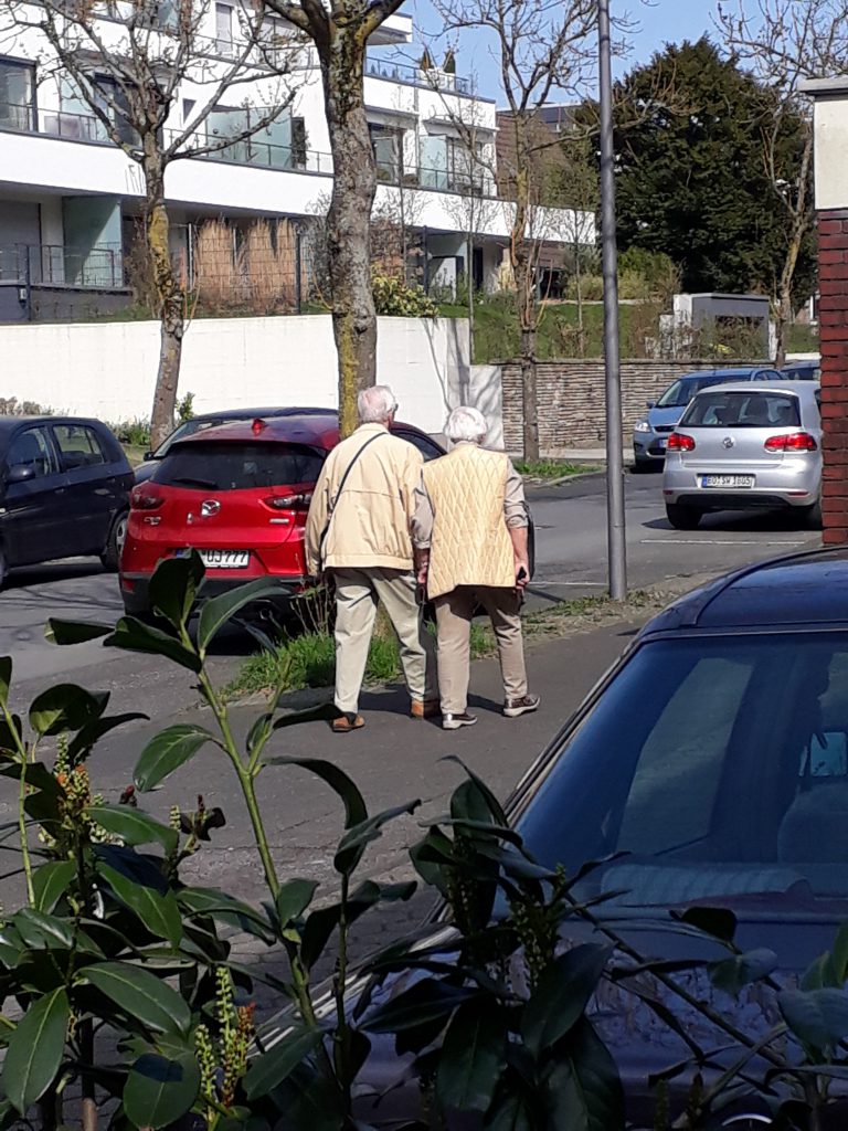 Älteres Paar in Beige spaziert einträchtig in gut situierter Wohngegend zwischen geparkten Autos.