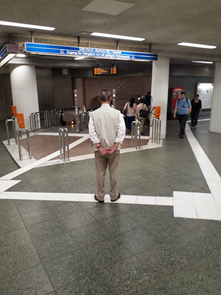 Mann in beiger Weste und dunkelbeiger Hose steht wartend in Unterführung vor den Rolltreppen mit dem Rücken zum Betrachter und verschränkt die Hände auf dem Rücken.