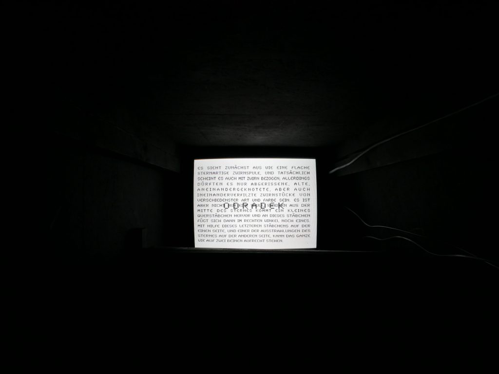 Schwarzer eng anmutender Raum mit Bildschirm an der hinteren Wand. Es steht dort schwarz auf weissem Grund u.a.: ODRADEK