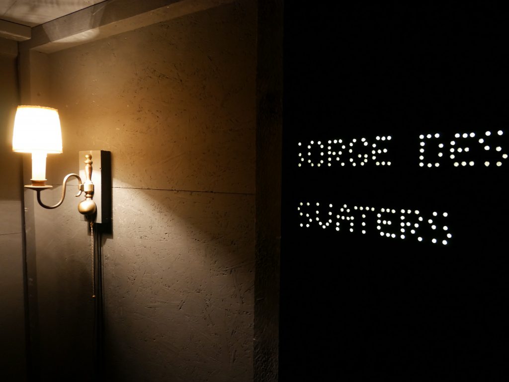 Eine Wandlampe beleuchtet eine hellbraune Wand. Vorne steht mit weissen Punkten auf schwarzem Grund:
SORGE DES SVATERS
