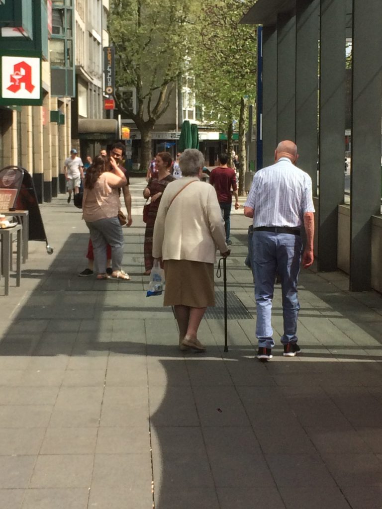 Paar in Fußgängerzone: Ältere Dame in dunkelbeigem Rock und hellbeiger Jacke, Herr in Bluejeans und weissem Hemd.