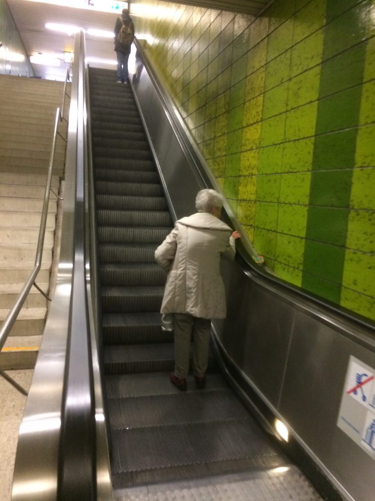 Dame in beigem Mantel steht auf Rolltreppe nach oben.