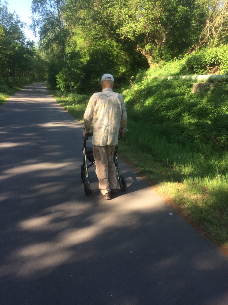 Mann in Beige läuft mit Rollator einen sommerlichen Spazierweg entlang.