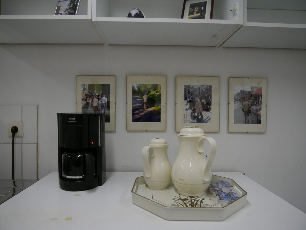 Ausstellungsansicht: 4 gerahmte Bilder, davor zwei cremefarbene Kaffeekannen und Kaffeemaschine.