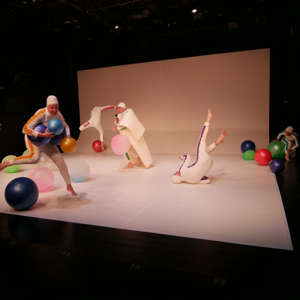 5 Performer in weißen enganliegenden Ganzkörperanzügen tanzen auf einer weissen Fläche mit bunten Bällen.