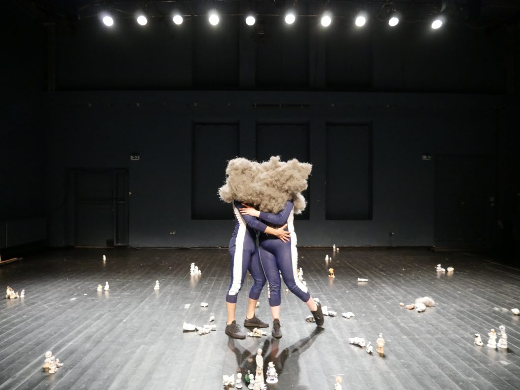 Bühnenansicht mit Porzellandfiguren und Karl und BechtOle, die sich umarmen, die Köpfe in einem dicken Staubknäuel versteckt. 