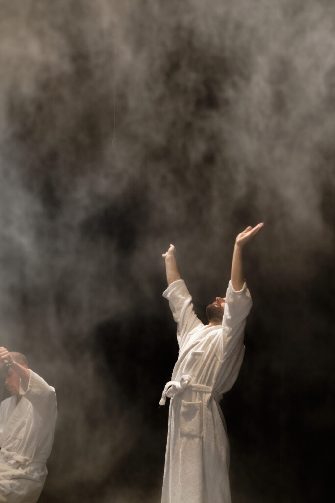Performer in weissem Bademantel hebt die Arme in ausladender Geste hoch Richtung aufsteigendem Nebel auf dunkler Bühne.