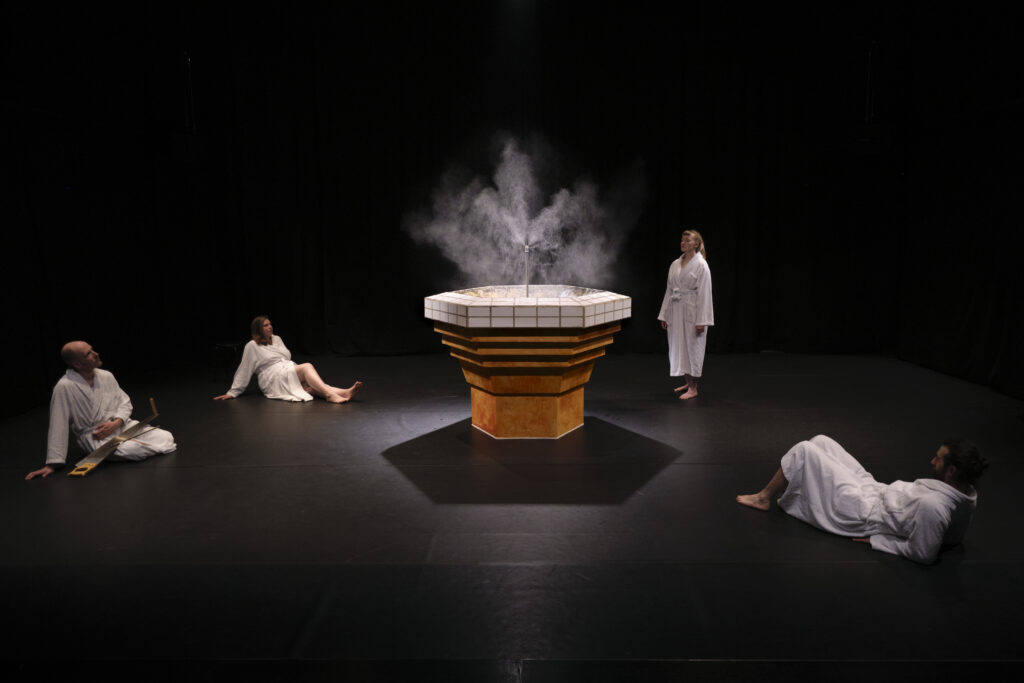 4 Performer auf dunkler Bühne in weissen Bademänteln, gruppiert um sechseckigen Brunnen aus dem Nebel aufsteigt 